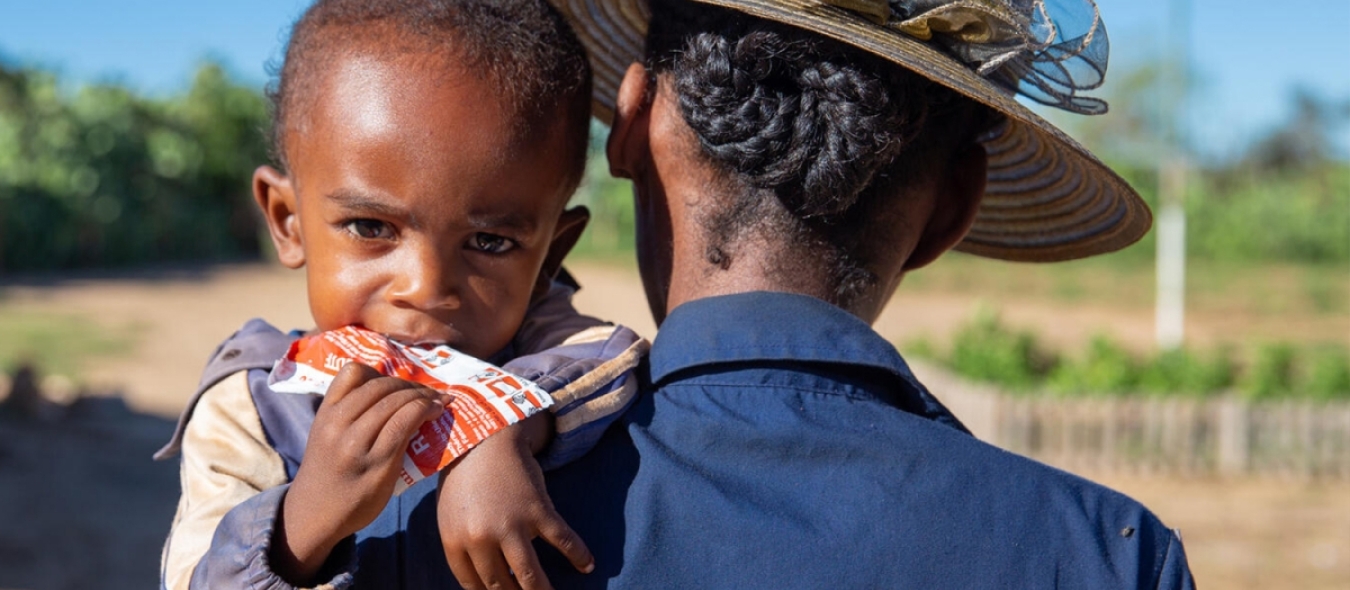 Mother carries her 2-year-old son while he eats ready-to-use therapeutic food (RUTF).