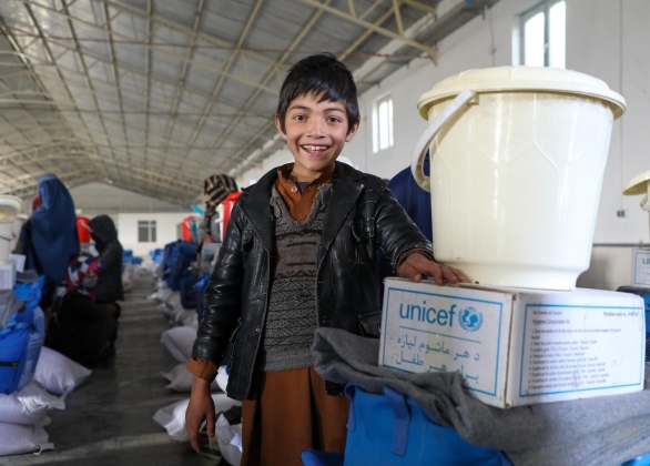 A smiling child stands next to a UNICEF winterization kit in Afghanistan.