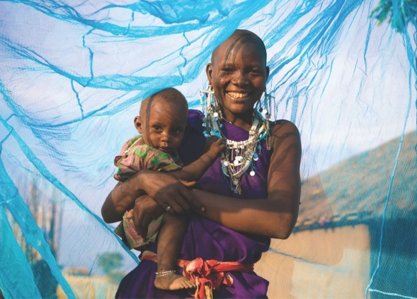A smiling mother holds her child, standing inside a bed net that protects them from mosquitoes.