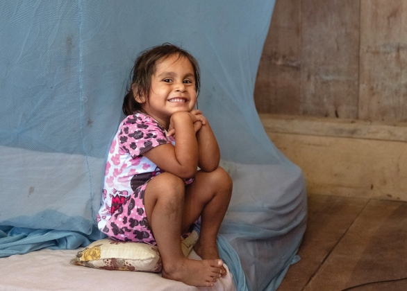 A smiling young girl sits on her bed which is covered with a mosquito net.